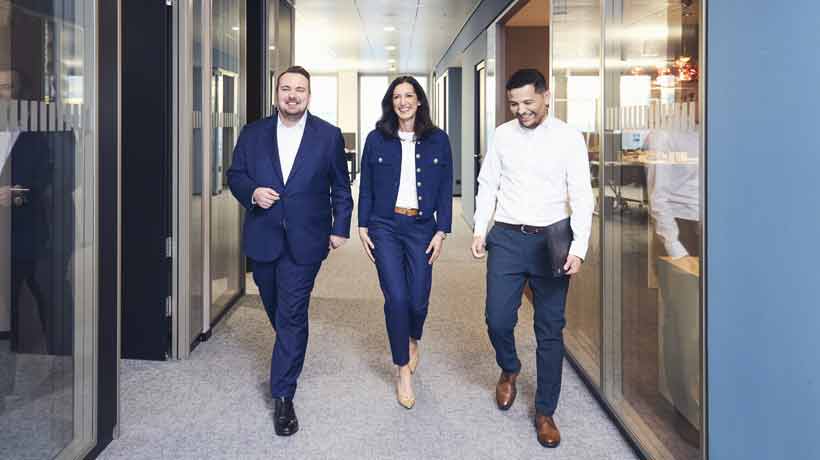 Three colleagues walk together in a modern office hallway.