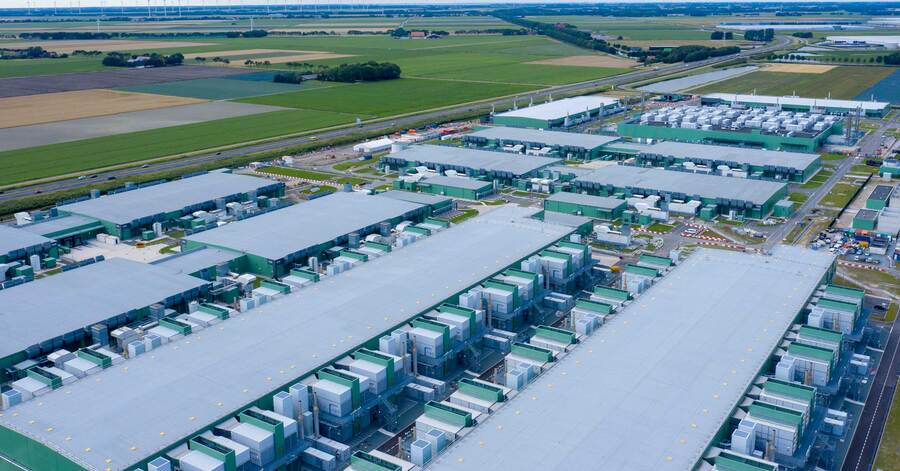 Aerial view of large data centre campus with multiple flat-roof buildings, cooling units and surrounding farmland.